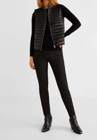 Schwarze gesteppte Weste mit Reißverschluss über einem enganliegenden schwarzen Pullover, kombiniert mit schwarzen, schlanken Hosen und Stiefeletten.