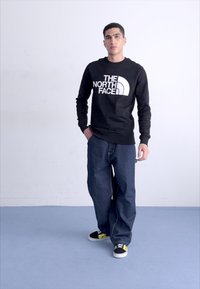 Sudadera negra con el logo blanco "THE NORTH FACE", combinada con jeans de corte holgado en azul y zapatillas negras con detalles amarillos.