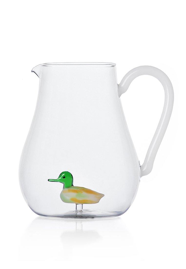Przezroczysty szklany dzbanek o zaokrąglonym kształcie z białym uchwytem, wewnątrz którego znajduje się kolorowa dekoracyjna figurka kaczki.