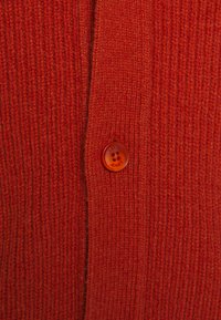 Cardigã vermelho de malha com um padrão de nervuras texturizado e um botão redondo laranja com o nome da marca em relevo.