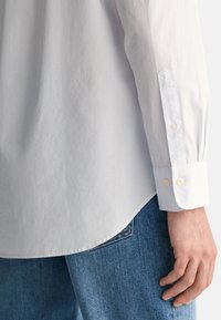 Camisa às riscas azul claro e branca com punhos de botão, feita de algodão, com bainha curva e textura suave. Usada com jeans azuis.