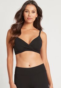 Zwarte crossover bralette met verstelbare bandjes; gladde stof, zachte textuur en een aansluitend ontwerp. Gecombineerd met zwarte high-waisted onderstukken.