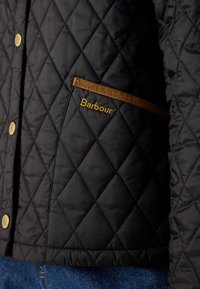 Jachetă neagră matlasată cu buzunar îmbrăcat în maro și logo-ul galben Barbour, purtată peste blugi albaștri.