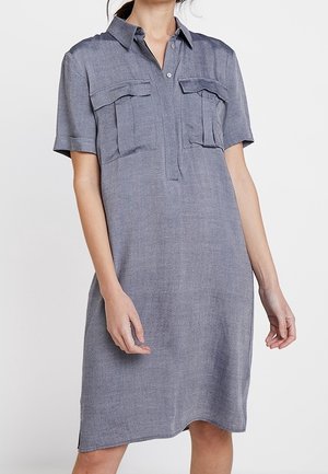 Kobieta ubrana w szarą sukienkę koszulową z krótkim rękawem, sięgającą do kolan, z dwoma przednimi kieszeniami na piersi i kołnierzykiem, stojąca na jednolitym tle.