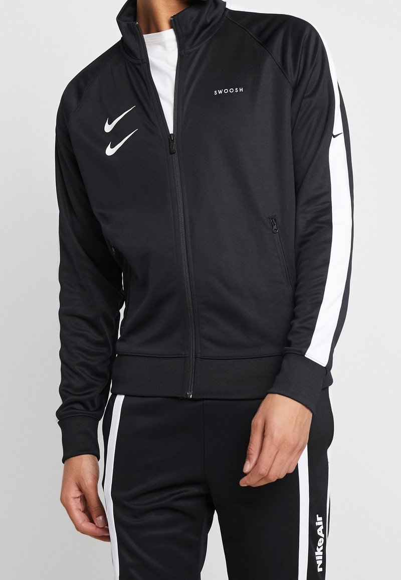Mies, joka pukeutuu mustaan Nike-trainingsutiin, jossa on valkoiset raidat, kaksi valkoista Nike-logoa ja "SWOOSH"-teksti takin rinnassa.