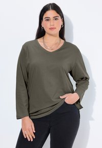Olive green long-sleeve shirt with a V-neck, made of a soft material, featuring a relaxed fit and side slit accents. Worn with dark jeans.