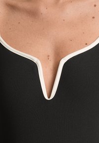 Fekete ujjatlan ruha, mély V-nyakkivágással, kontrasztos fehér díszítéssel a nyakkivágás körül. Sima textúra, minimalista dizájn.