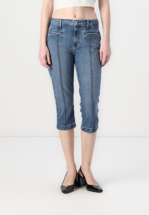 Femme portant un jean bleu cropped taille mi-haute avec des coutures avant et des escarpins slingback en cuir verni noir, debout devant un fond blanc.