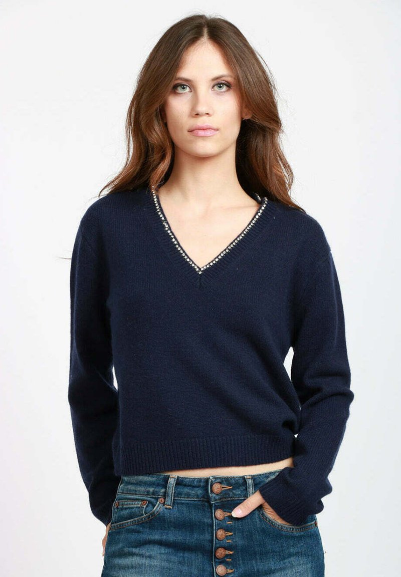 Giovane donna con lunghi capelli castani, che indossa un maglione blu navy a V con applicazioni di strass e jeans blu a vita alta con bottoni.