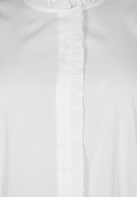 Vit bomullsblus med en central volangdetalj längs knappraden och en slät yta med rund halsringning.