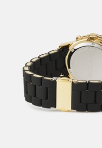 Correa de reloj de silicona negra con acentos metálicos en tono dorado y hebilla. Esfera del reloj redonda visible con una parte posterior suave y reflectante.