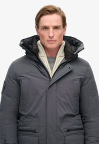 Homme portant une veste d'hiver grise avec capuche sur un pull beige, faisant face à l'objectif sur un fond blanc uni.