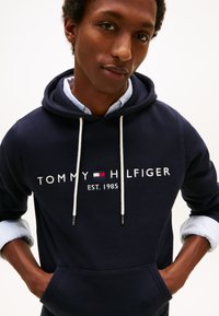 Sudadera con capucha azul marino con cordones blancos, con el logotipo "TOMMY HILFIGER EST. 1985" en blanco y detalles en rojo. Tela suave, bolsillo frontal.