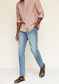 Camisa de rayas rojas y blancas de manga larga, jeans azul claro y sandalias marrón oscuro. La camisa tiene cierre de botones y mangas arremangadas.