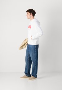 Hanorac alb cu logo-uri roșii, blugi din denim albastru și adidași bej. Modelul ține o jachetă bej. Design simplu și croială lejeră.