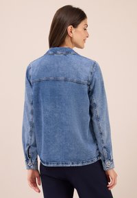 Vrouw met lang bruin haar draagt een middelblauwe denimjas en donkere broek, staande tegen een effen beige achtergrond, zijwaarts gekeerd.