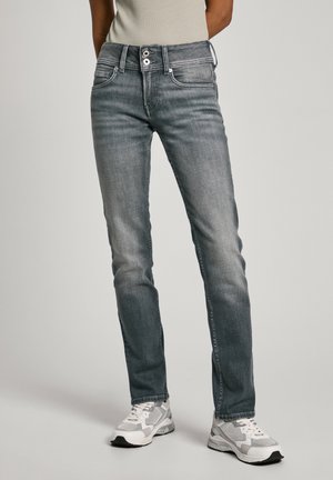 Personne portant un jean gris slim à taille double bouton et des baskets blanc-gris, debout devant un fond neutre.