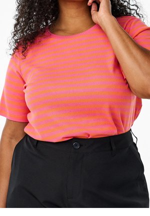 Kvinde iført en knaldpink og orange stribet kortærmet skjorte stoppet ind i sorte bukser med højtalje, med hånden tæt på ansigtet.