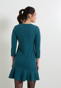Vestido en teal con textura, mangas tres cuartos, cuerpo ajustado y un dobladillo acampanado con volantes. Escote simple que resalta un diseño estructurado.