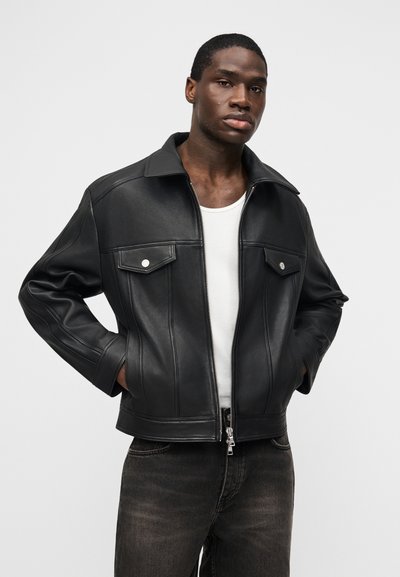 Άνδρας μοντέλο με μαύρο δερμάτινο μπουφάν πάνω από λευκή αμάνικη μπλούζα και σκούρο τζιν, όρθιος με τα χέρια στις τσέπες του μπουφάν σε λευκό φόντο.