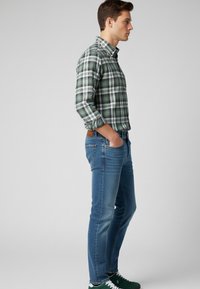 Camisa a cuadros verde y blanca con cuello tipo polo, combinada con jeans azul slim-fit y zapatillas verdes. Vista lateral, fondo neutro.