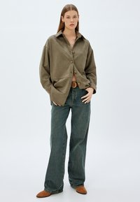 Camisa de botones verde oliva con un corte relajado, combinada con jeans azul de cintura alta y zuecos de ante marrón. Textura suave, con detalles mínimos.