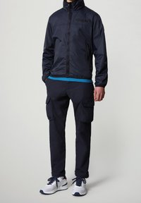 Giacca a vento navy, con zip frontale, colletto alto e dettagli con logo; abbinata a pantaloni cargo navy e scarpe sportive bianche con accenti navy.