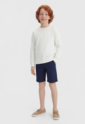 Boy with red hair wearing a white sweatshirt, navy blue shorts, and tan boat shoes standing and smiling against a plain background.