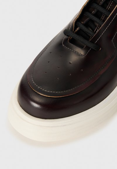 Sneaker en cuir marron foncé avec un bout arrondi, une surface texturée, des lacets noirs contrastants, et une épaisse semelle blanche avec des rainures horizontales.