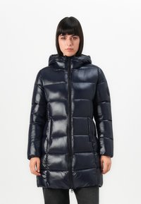 Vrouw in een glanzende zwarte gewatteerde pufferjack met capuchon, met een rits aan de voorkant en zakken, staande tegen een witte achtergrond.