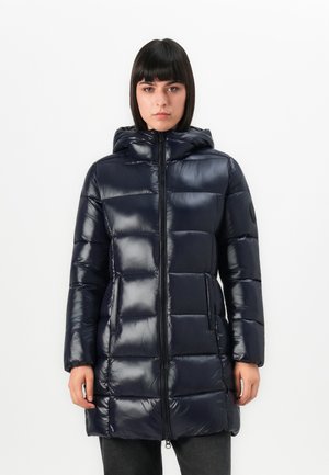 Vrouw in een glanzende zwarte gewatteerde pufferjack met capuchon, met een rits aan de voorkant en zakken, staande tegen een witte achtergrond.