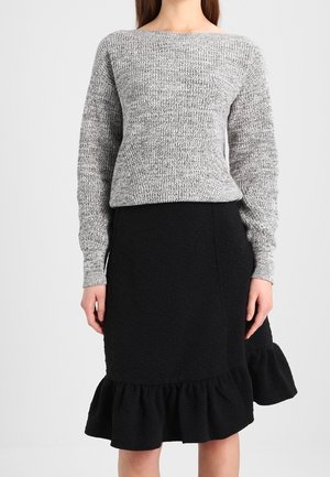 Femme portant un pull gris en maille à manches longues et une jupe noire longueur genou avec un ourlet à volants.