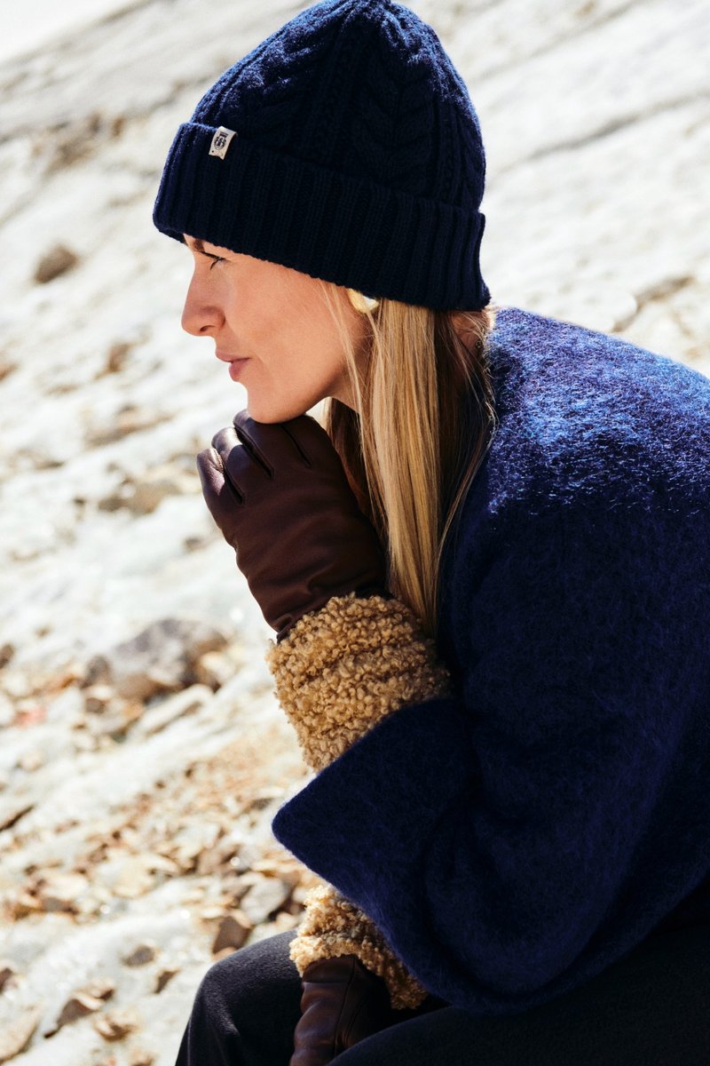 Navy knit beanie with cable pattern, brown leather gloves, and a textured cream wool-collared outerwear. Outdoor setting with rocky ground.