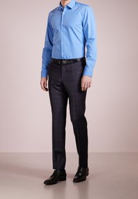 Lichtblauw overhemd met lange mouwen en knopen, gecombineerd met donker geruite op maat gemaakte broek en zwarte nette schoenen, aangevuld met een zwarte riem.