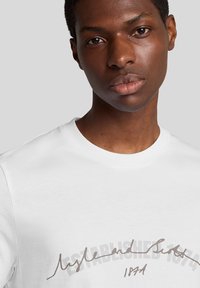 Hvid bomulds t-shirt med rund halsudskæring og et trykt skriftligt design i brun, med teksten "Etableret 1974" tydeligt fremhævet.