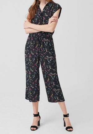 Femme portant une combinaison noire à manches courtes fleurie avec des jambes larges raccourcies et des sandales noires à talons blocs avec bride à la cheville, debout, les bras croisés.