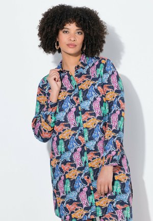 Kvinde med krøllet hår iført en langærmet knap-op kjole med farverige gepardmønstre på en mørk baggrund.