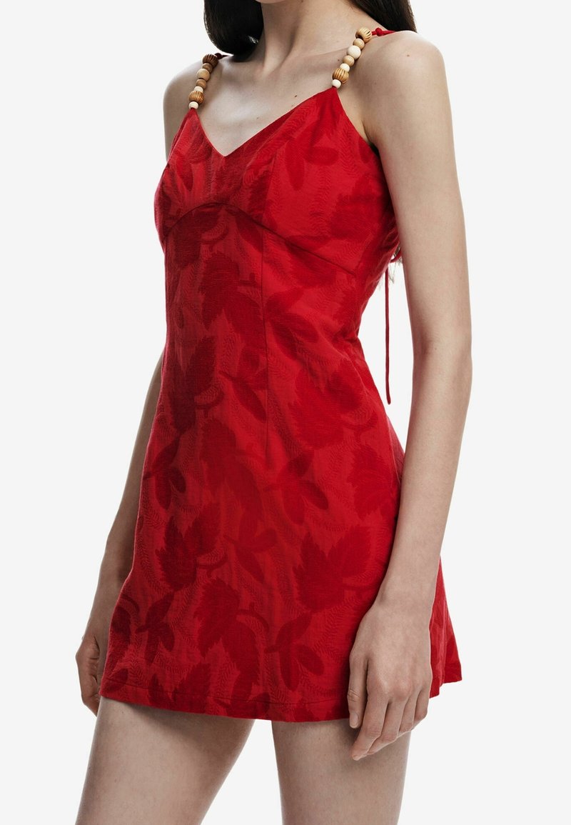 Femme portant une robe rouge sans manches avec des motifs floraux et des bretelles en perles de bois, debout avec un bras détendu le long du corps.