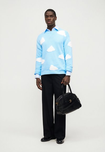 Άντρας με ανοιχτό μπλε πουλόβερ με μοτίβα λευκών σύννεφων, μαύρο παντελόνι, κρατώντας μαύρη δερμάτινη τσάντα, στέκεται σε λευκό φόντο.