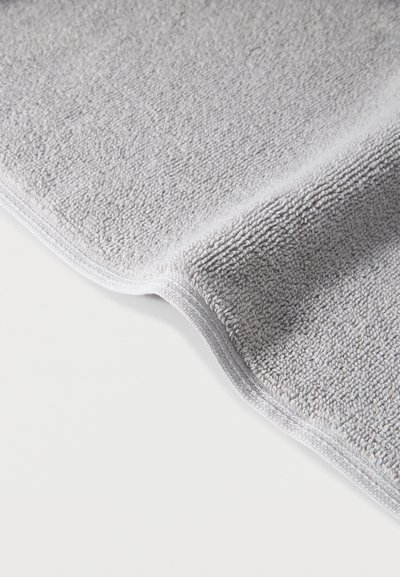 Απαλή, διπλωμένη πετσέτα από ανοιχτό γκρι περκάλι με ορατούς βρόχους και ραμμένες άκρες πάνω σε επίπεδη λευκή επιφάνεια.