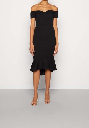 Robe noire moulante épaules dénudées avec manches courtes et ourlet à volants, portée avec des talons ouverts transparents sur un fond clair.