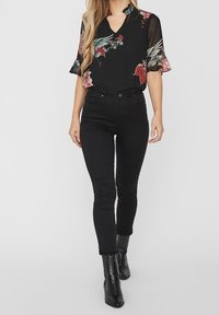 Blusa nera a fiori con maniche corte a volant, abbinata a jeans skinny neri e stivaletti. La blusa presenta motivi vivaci in rosa e verde.