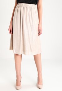 Wanita mengenakan rok plisket beige sepanjang lutut dipadukan dengan atasan hitam dan sepatu hak tinggi beige, berdiri di depan latar belakang putih.