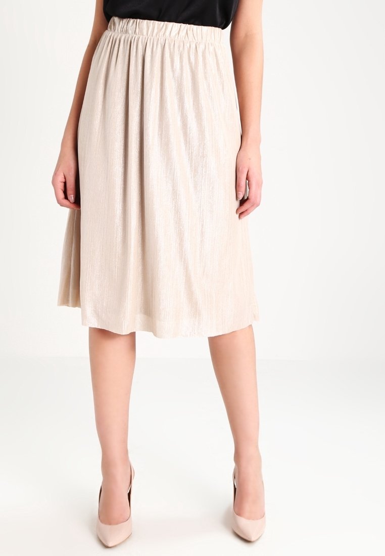 Wanita mengenakan rok plisket beige sepanjang lutut dipadukan dengan atasan hitam dan sepatu hak tinggi beige, berdiri di depan latar belakang putih.