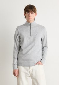 Pulover gri cu fermoar, dintr-un material texturat, cu guler înalt și un mic logo negru. Mâneci lungi și tiv cu nervuri. Purtați peste pantaloni de culoare deschisă.