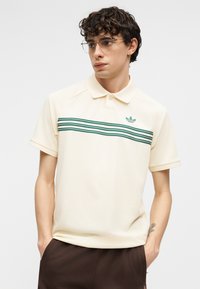 Junger Mann mit lockigem Haar, der eine Brille mit klarem Rahmen und ein cremefarbenes Poloshirt mit grünen Streifen trägt, Hände in den Taschen einer dunkelbraunen Hose.