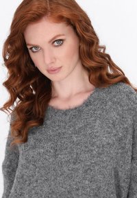 Grauer Strickpullover mit weichem Textil, weitem Ausschnitt und langen Ärmeln. Der Stoff wirkt flauschig und sorgt für Wärme und Komfort.
