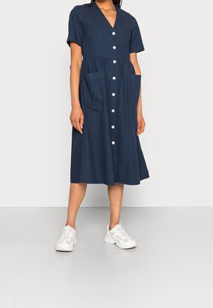 Kvinde iført en mørkeblå midi-kjole med knapper foran, korte ærmer og forlommer, kombineret med hvide kraftige sneakers.