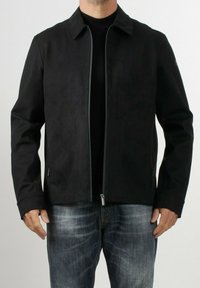 Giacca nera con texture liscia, collo a camicia, chiusura frontale con zip e tasche laterali, indossata sopra una maglietta nera e jeans blu.