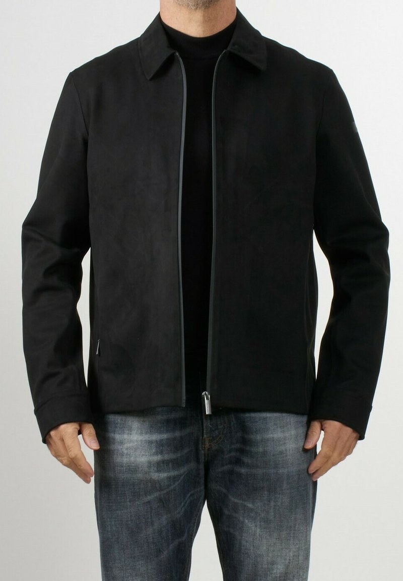 Giacca nera con texture liscia, collo a camicia, chiusura frontale con zip e tasche laterali, indossata sopra una maglietta nera e jeans blu.
